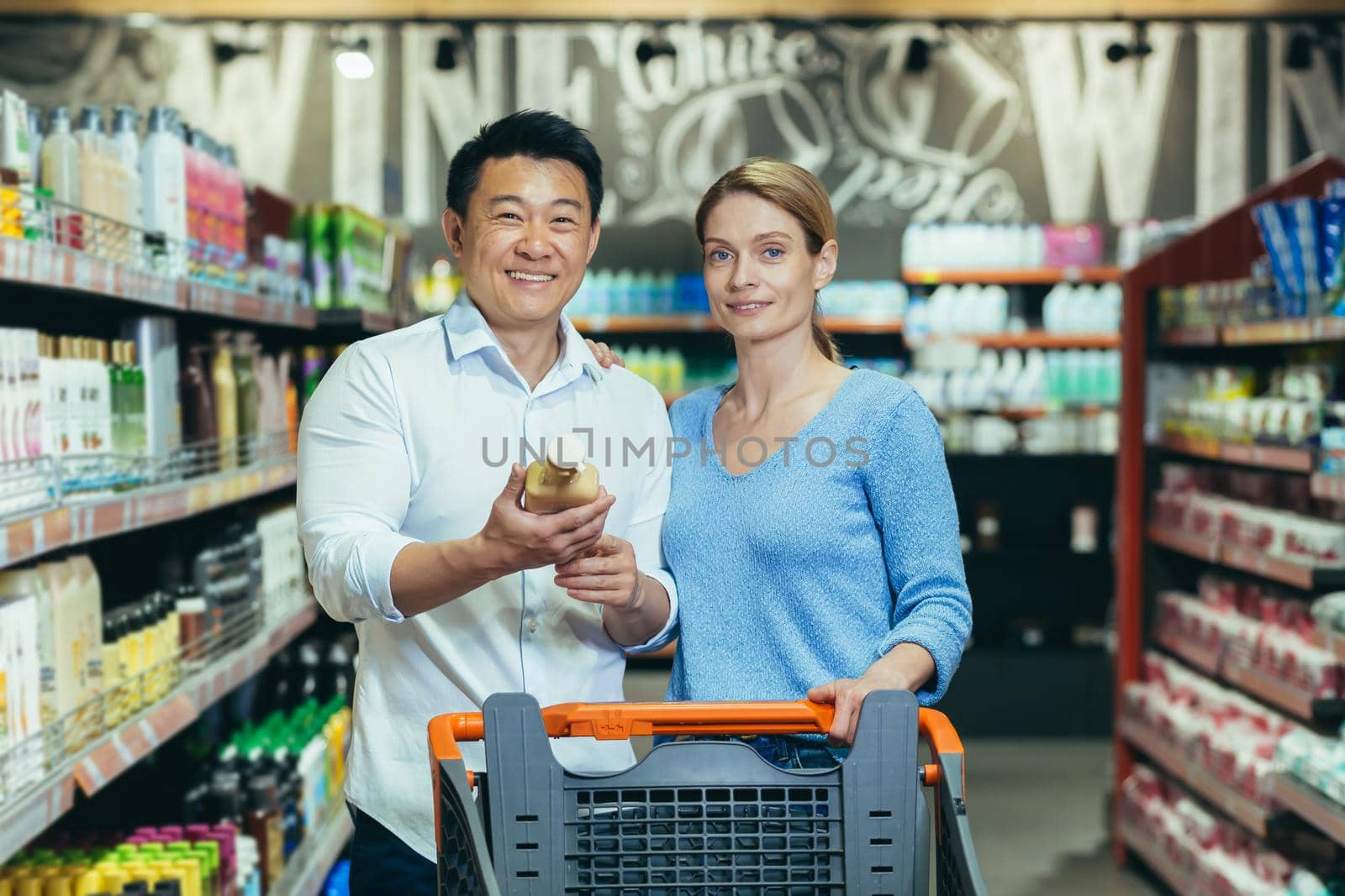A young international couple, a woman and an Asian man are walking between the rows with a trolley in the supermarket. The man is holding a bottle of shampoo in his hands, looking at the camera, smiling.