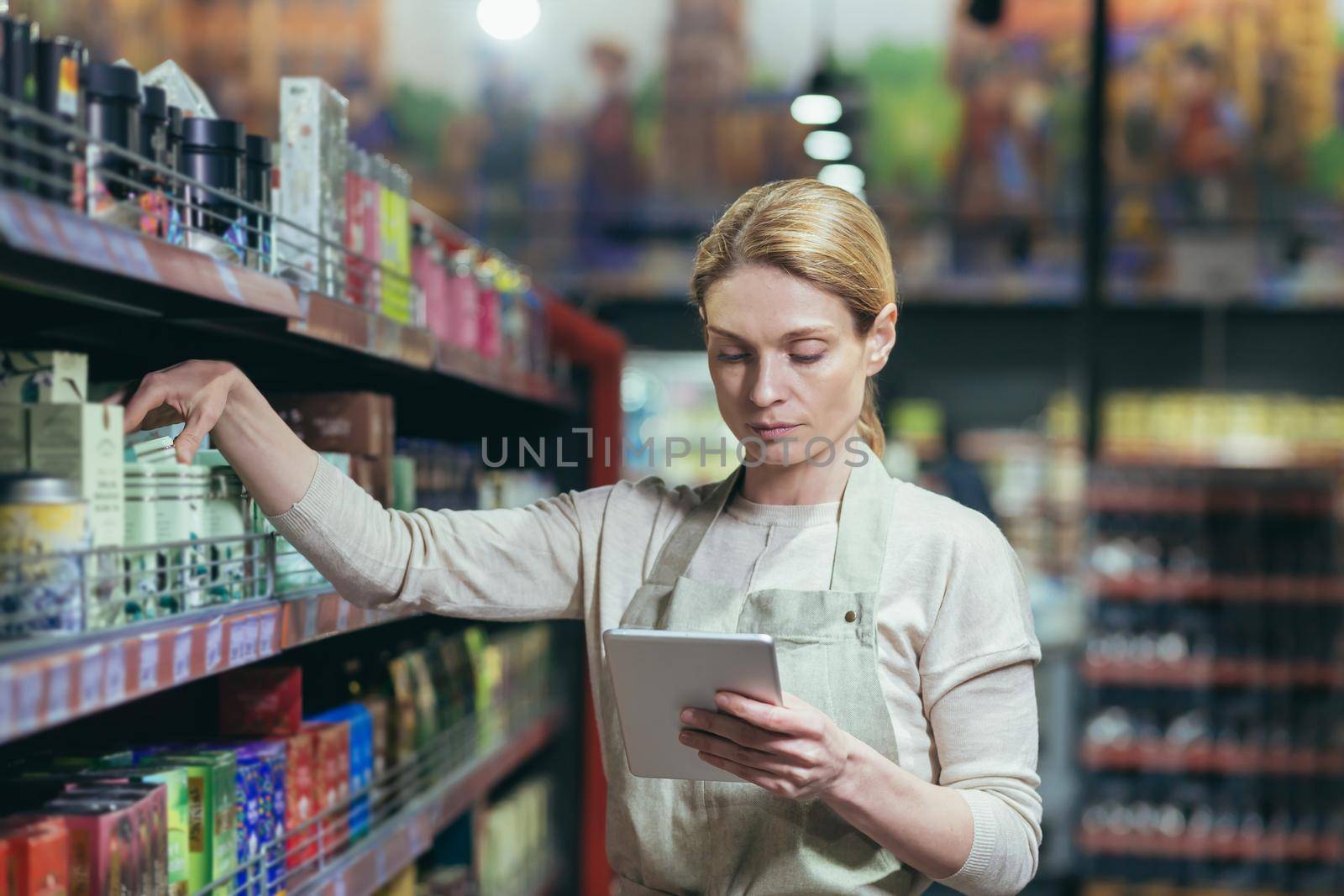 A beautiful young worker in an apron is taking inventory in a supermarket, store. He stands with a notebook in his hands and takes a product from the shelf.