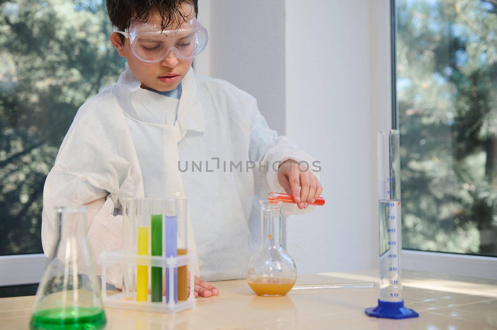 Handsome smart Caucasian schoolboy in a white lab coat and protective eyewear, experimenting in chemistry class, pouring liquid from test tube into a lab flask, in the science class.Education concept