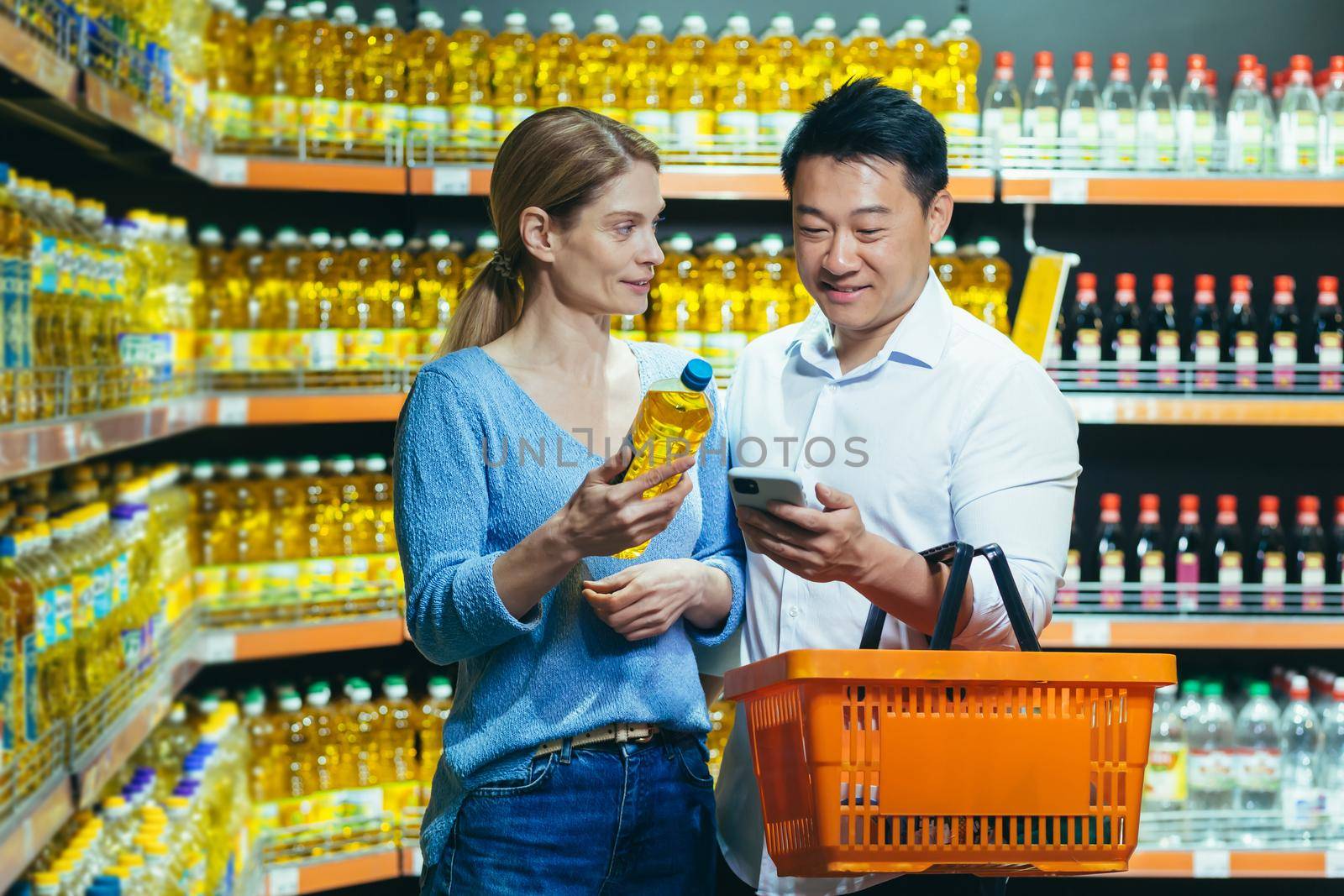 Young international family, Woman and asian man choose oil in supermarket. They hold a bottle of oil, a basket and a phone. They are looking for the brand, composition of the oil in the search engine.