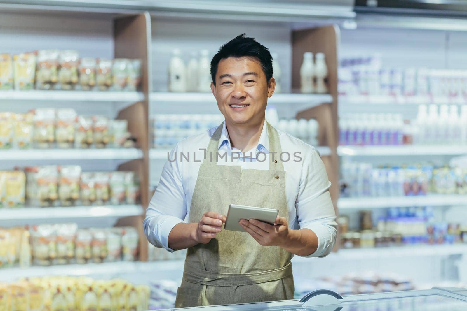 Portrait of a young handsome Asian man, shop owner, a supermarket worker. Standing in an apron in the dairy department of the store with a notebook in his hands, looking at the camera, smiling.