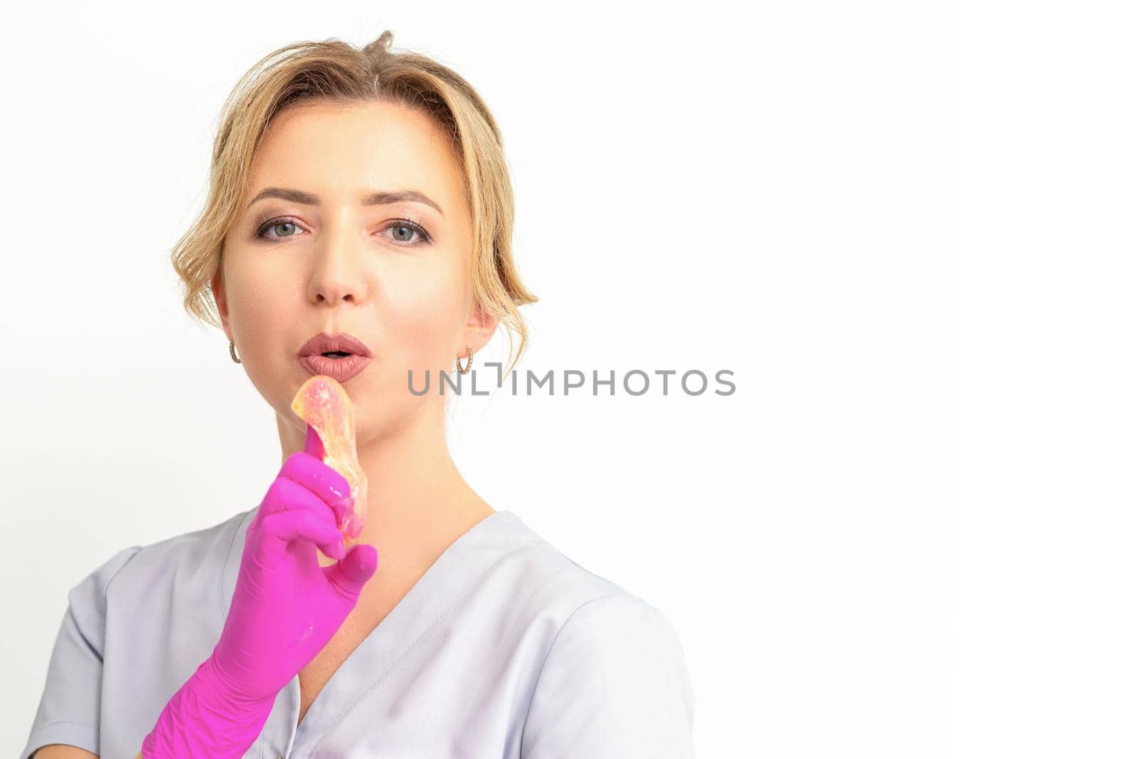 Young caucasian female beautician holding wax paste on forefinger blowing on him wearing pink gloves against a white background. Epilation concept