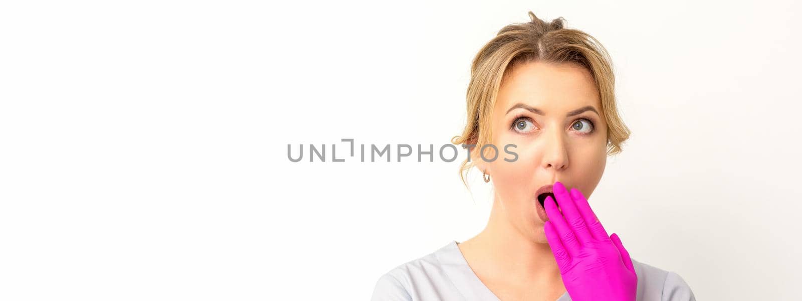 Portrait of a young female caucasian doctor or nurse is shocked covering her mouth with her pink gloved hands against a white background. by okskukuruza