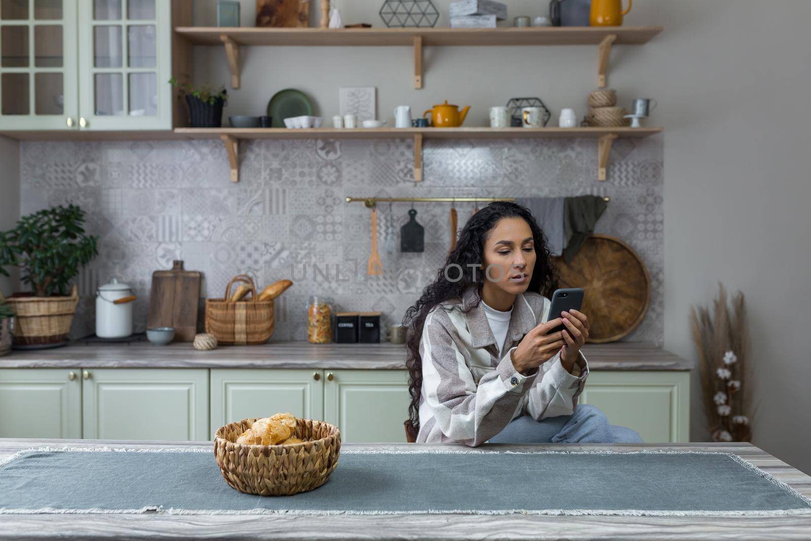 Serious and thoughtful Latin American woman in the kitchen uses the phone, woman thoughtfully reads a message from an online application. by voronaman