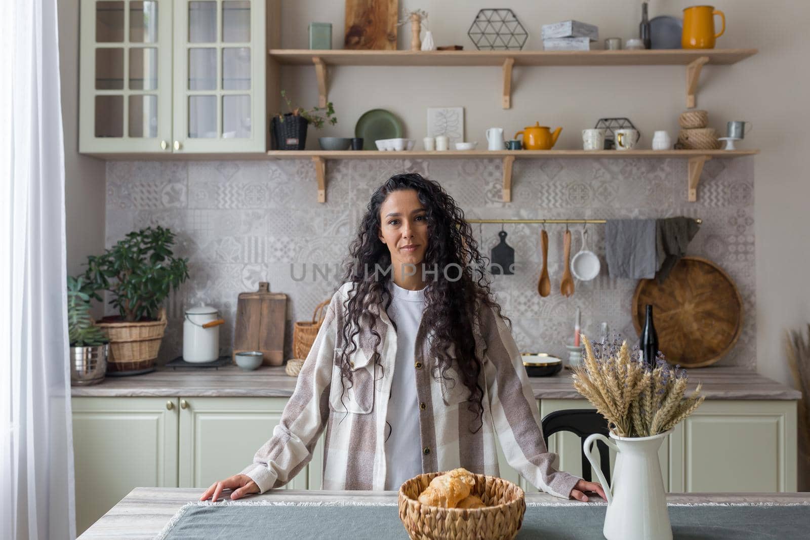 Portrait of happy hispanic woman at home in kitchen, woman with curly hair smiling and looking at camera.