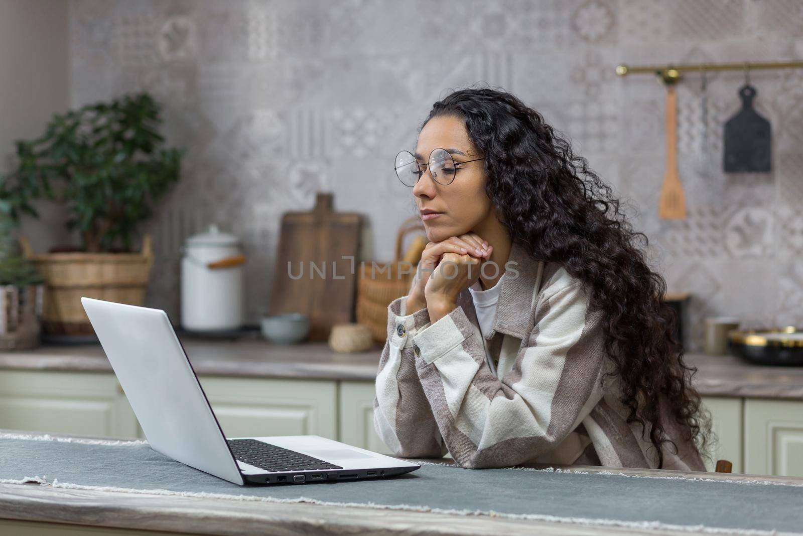Hispanic woman thinking at home sitting in kitchen working on laptop, freelancer in glasses and curly hair, businesswoman working and learning online remotely.