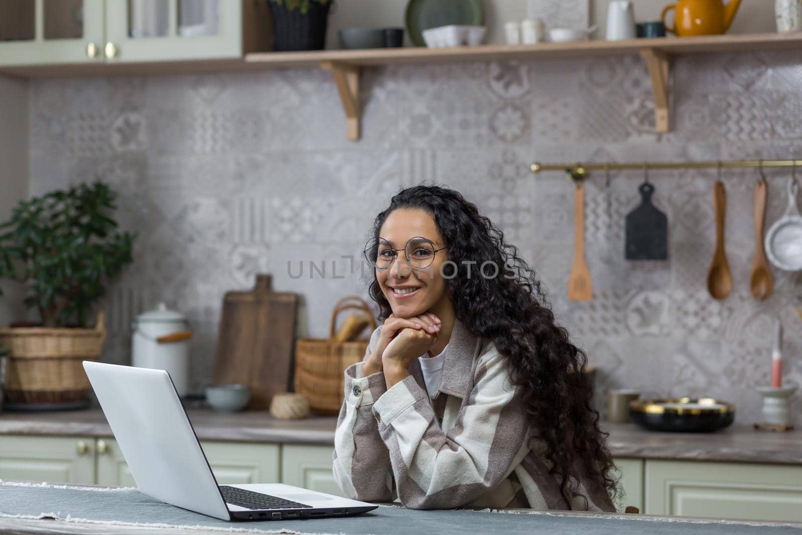 Portrait of young beautiful hispanic woman at home, female freelancer working remotely using laptop, looking at camera and smiling in glasses and curly hair in kitchen