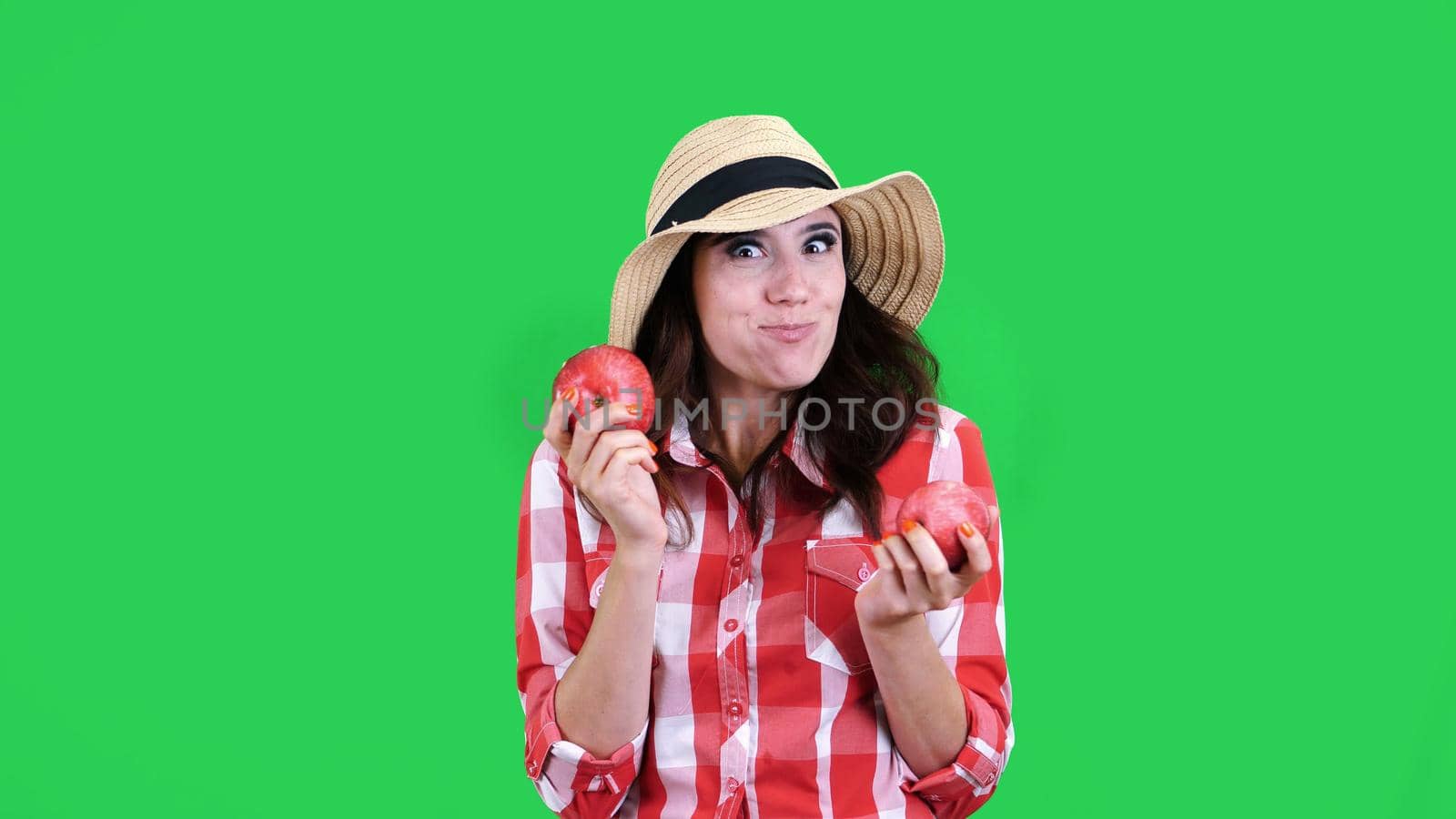 funny, smiling female farmer in checkered shirt and hat, holding in hands two large sour apples, eats, bites them, wrinkles her face, on green background in studio. Healthy food to your table. High quality photo