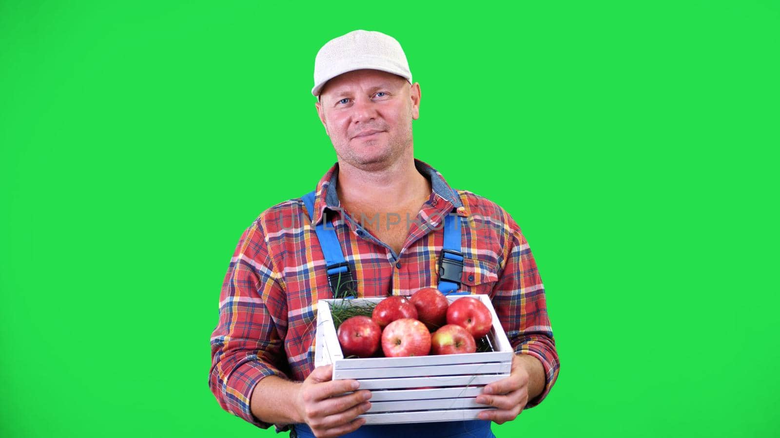 portrait of handsome male farmer holding a wooden box with red ripe organic apples, smiling. on green background in studio. Healthy food to your table. High quality photo