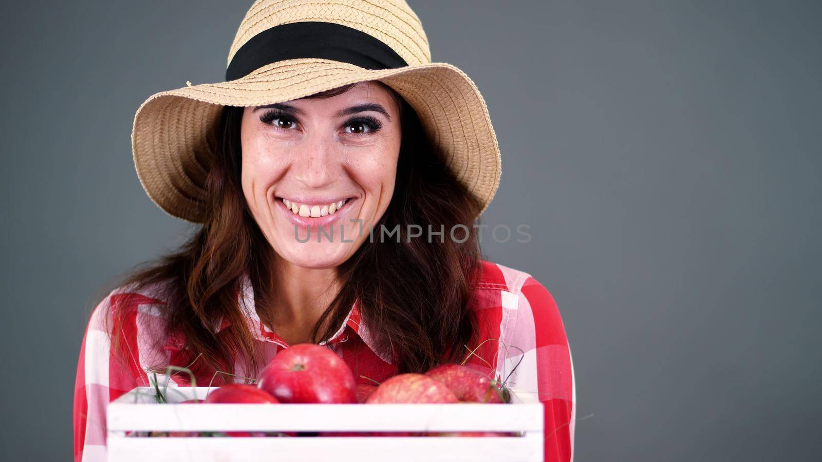 portrait of beautiful smiling female farmer in plaid shirt, gloves and hat holding white wooden box with red ripe organic apples, on gray background, in studio,. High quality photo