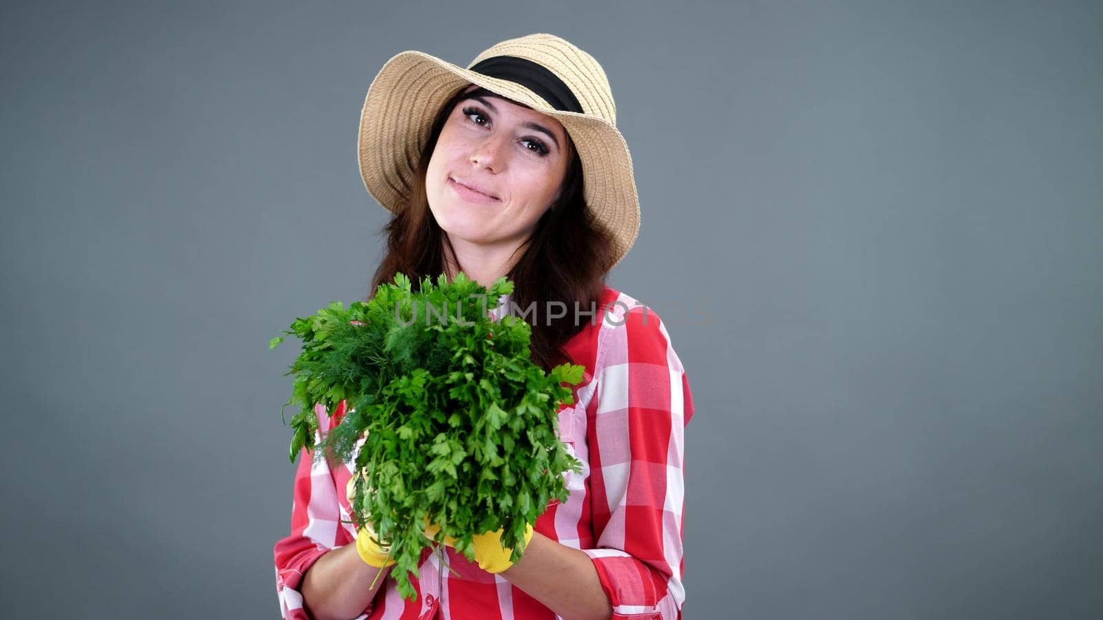 portrait of smiling female farmer in plaid shirt, gloves and hat holding, smells a bunch of fresh green parsley, greens, on gray background, in studio. Healthy food to your table. High quality photo