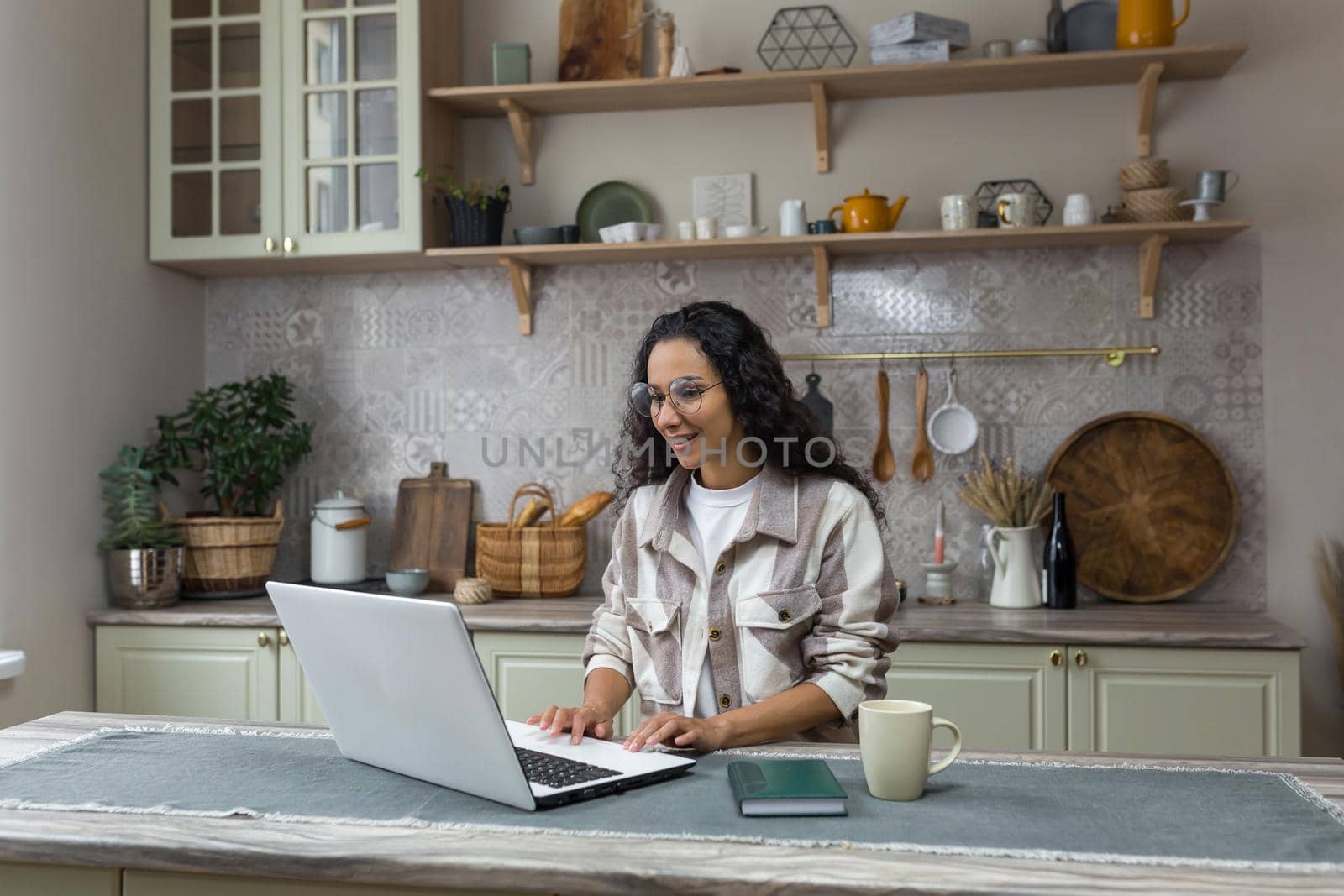 Online learning at home, young latin american woman with curly hair and glasses studying remotely at home using laptop, female student sitting in the kitchen at the table.