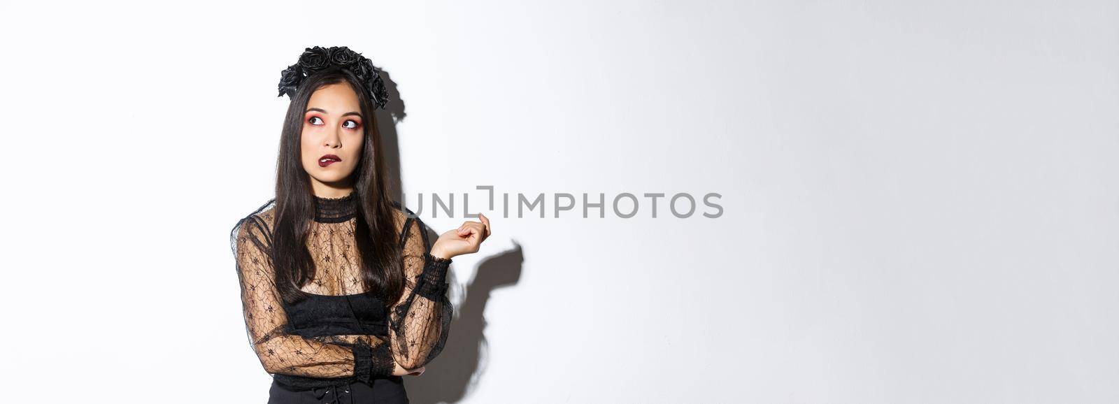 Thoughtful beautiful asian woman in witch costume biting lip and looking upper left corner, daydreaming while standing in halloween widow dress, standing over white background.