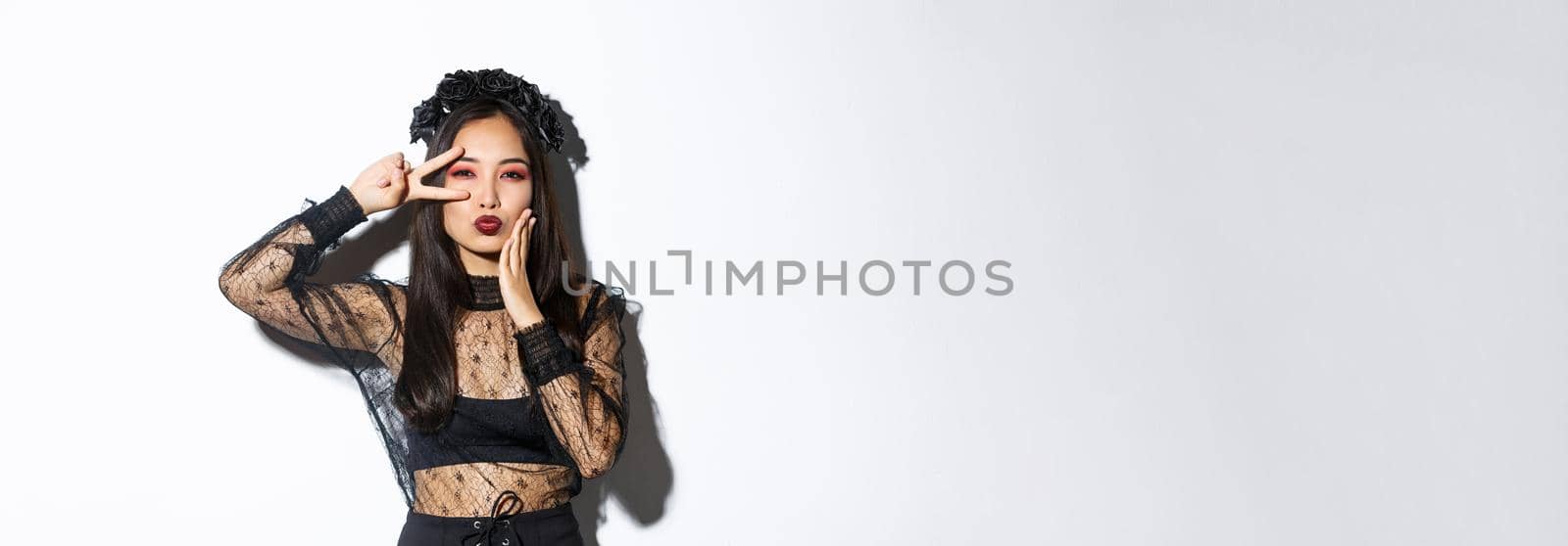 Image of beautiful asian woman in gothic lace dress and black wreath posing on halloween banner, showing peace gesture and pouting, standing over white background.