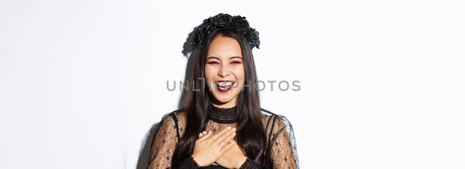 Image of evil witch press hands to chest and laughing, girl celebrating halloween and having fun, standing over white background.