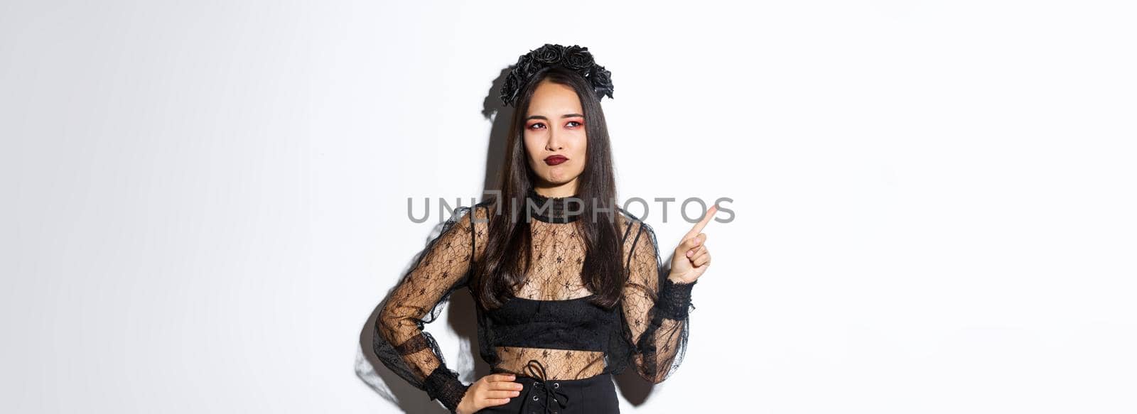 Unamused and skeptical asian beautiful woman in witch dress, looking at upper left corner with displeased smirk, standing over white background, showing logo or promo banner.