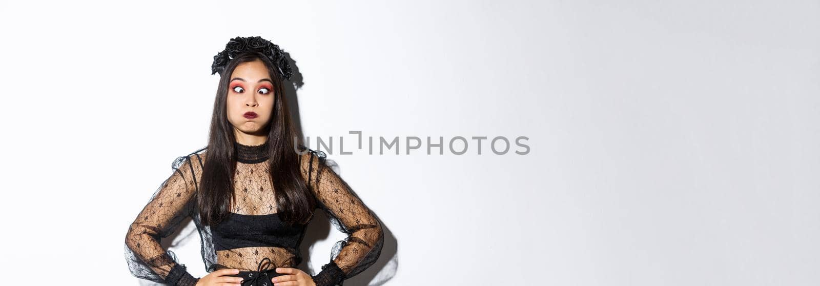 Portrait of funny and carefree asian girl having fun on halloween, making faces and holding breath, standing in gothic witch costume over white background by Benzoix