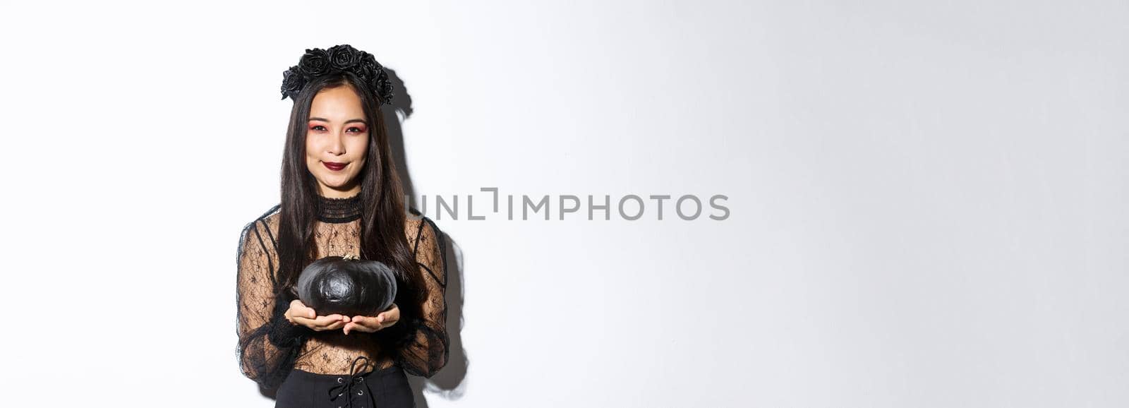 Smiling devious asian girl in witch costume, celebrating halloween, holding black pumpkin, standing over white background.