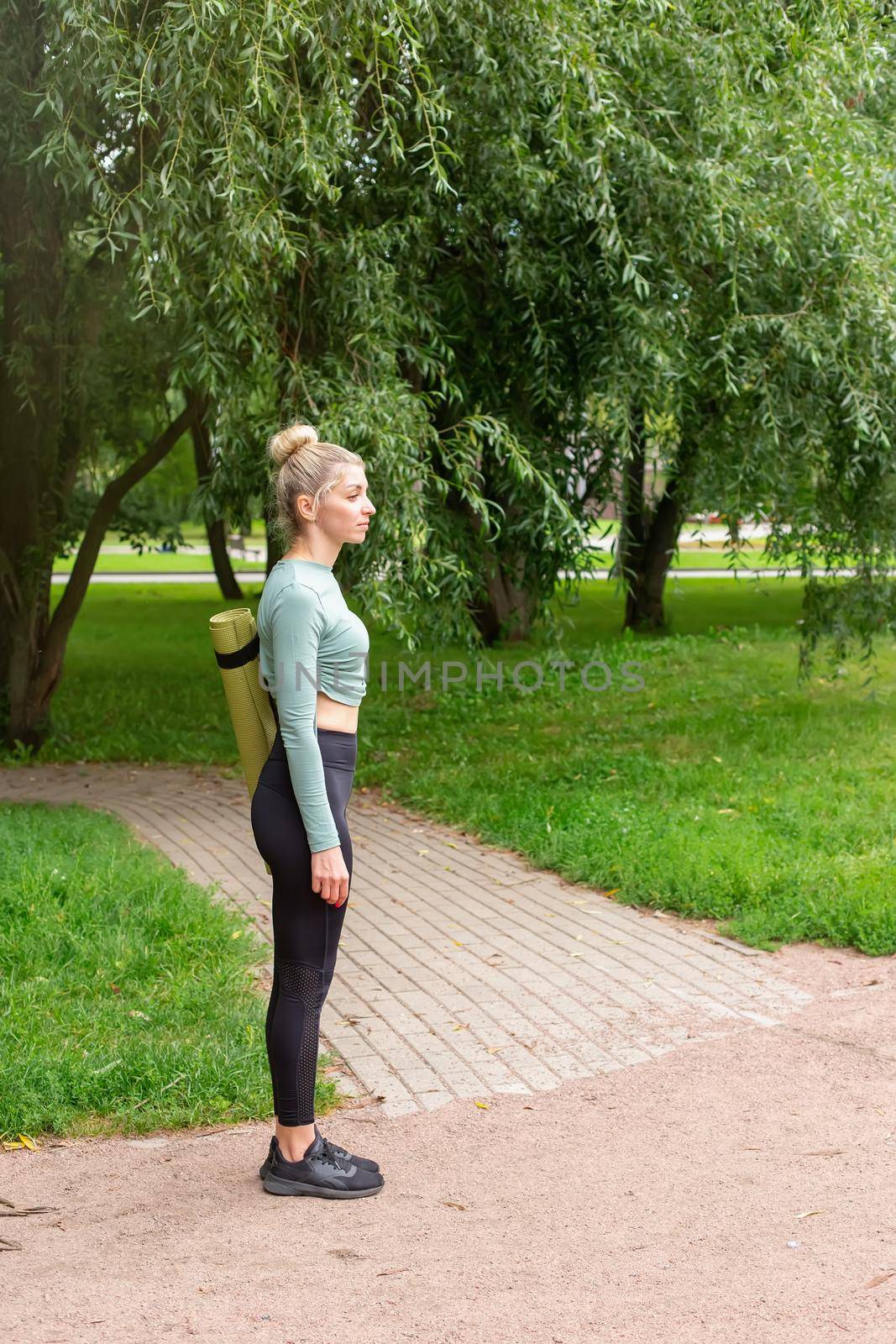 A slender woman in sportswear, standing in summer in park, with a green gym mat for sports. by Zakharova