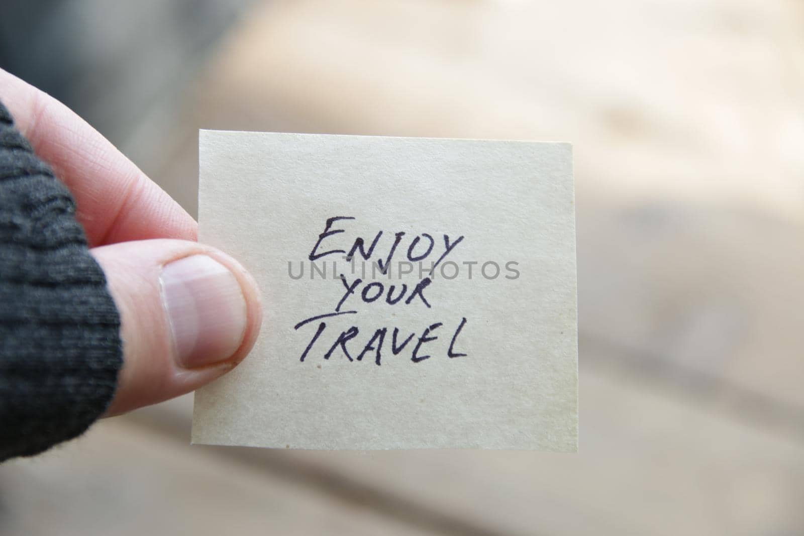 A hand holds a tag with the inscriptionEnjoy your travel on a blurred background.