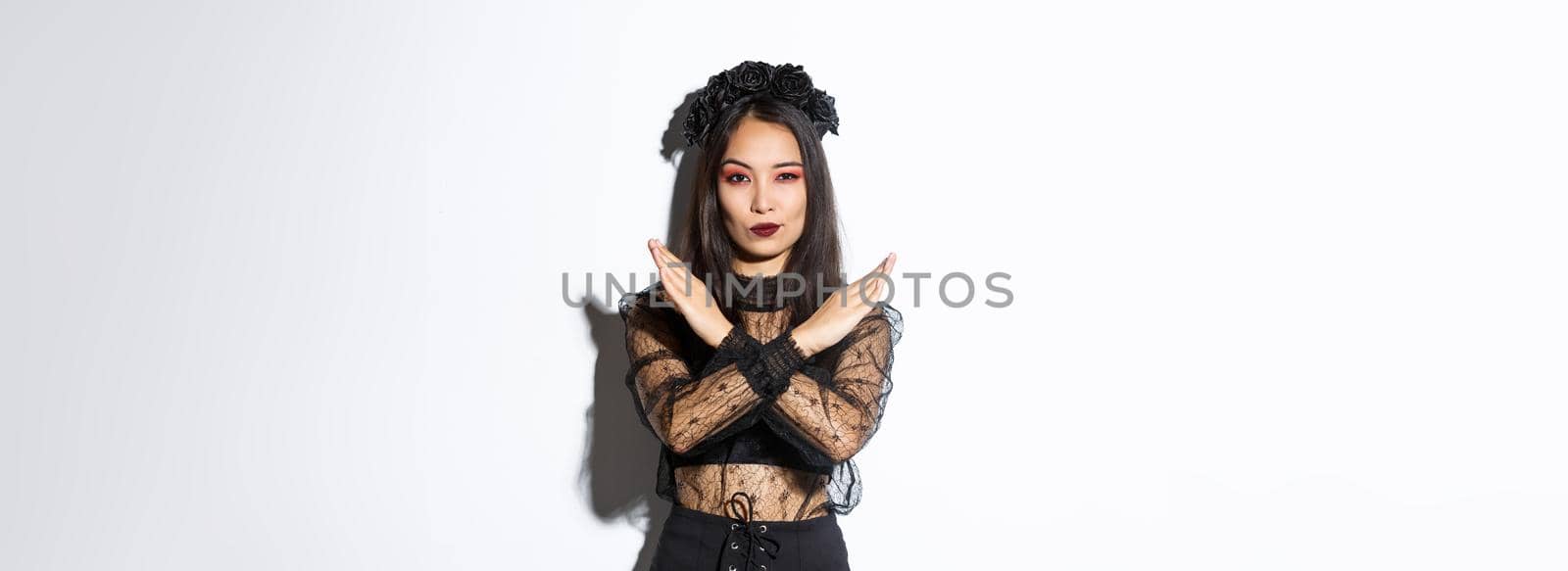 Confident beautiful woman in black gothic dress showing cross gesture, disapprove and stop something bad, disagree with someone about halloween, standing over white background by Benzoix