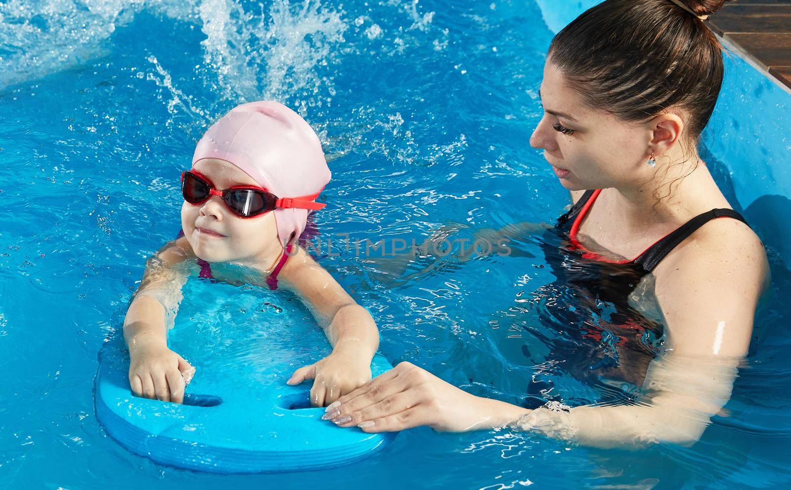 Little girl learning to swim in indoor pool with pool board and trainer by Mariakray