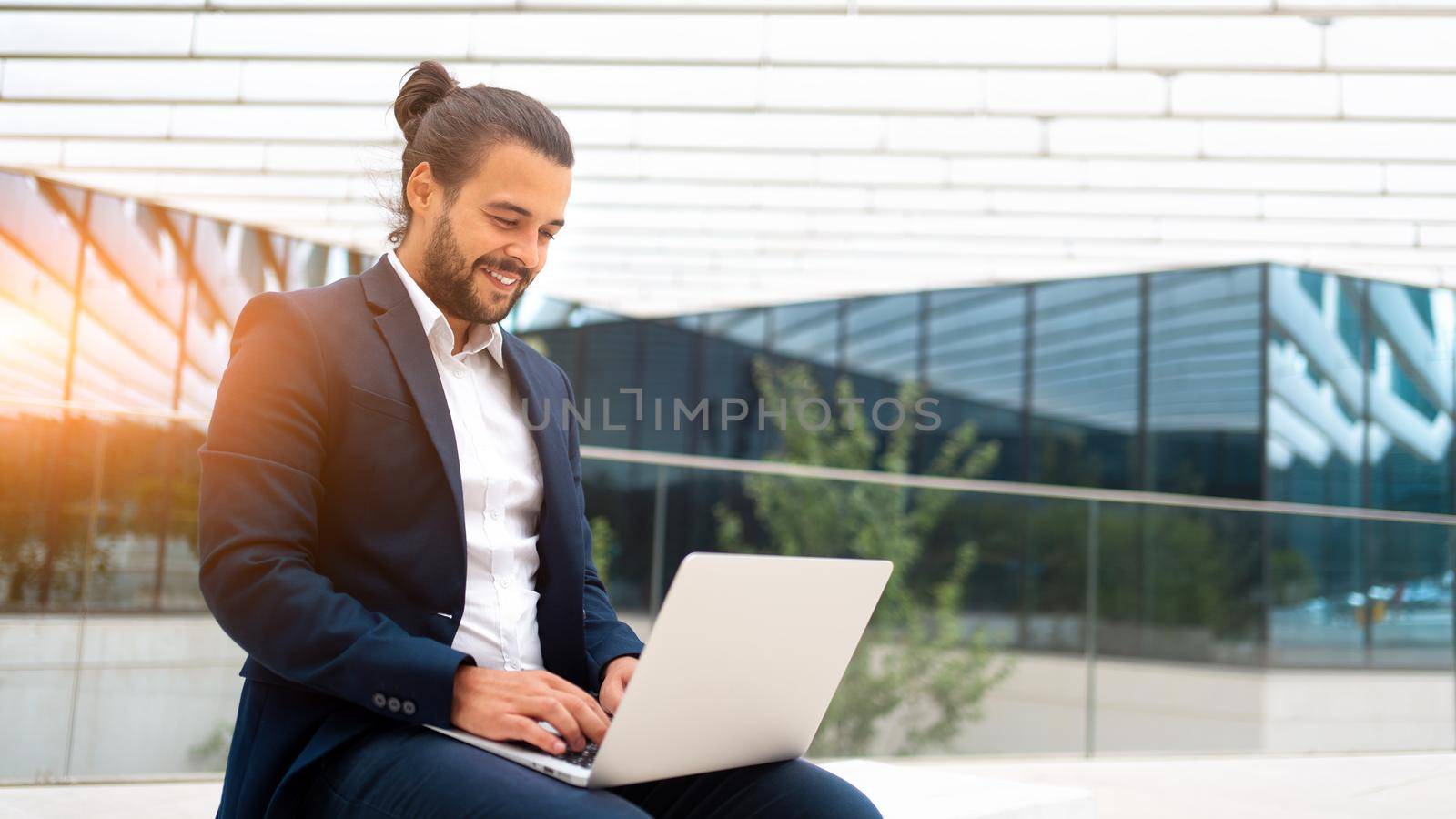 Business on the Go Businessman Working on his Laptop on the Street near office building. Handsome hispanic business person sitting use laptop. Confident male manager working outdoor. Banner size.