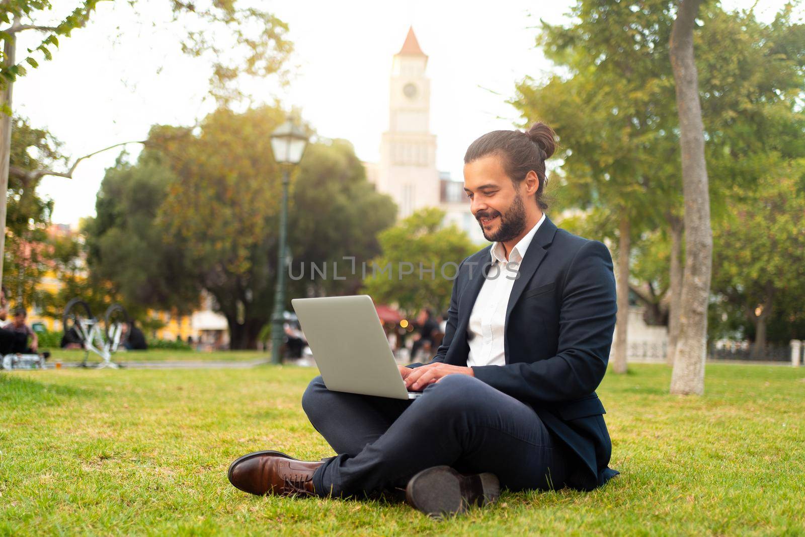Handsome hispanic male businessman sitting lotus position in public park use laptop. Remote work concept. Stylish european man in suit relax in park summer day working on computer. Technology.