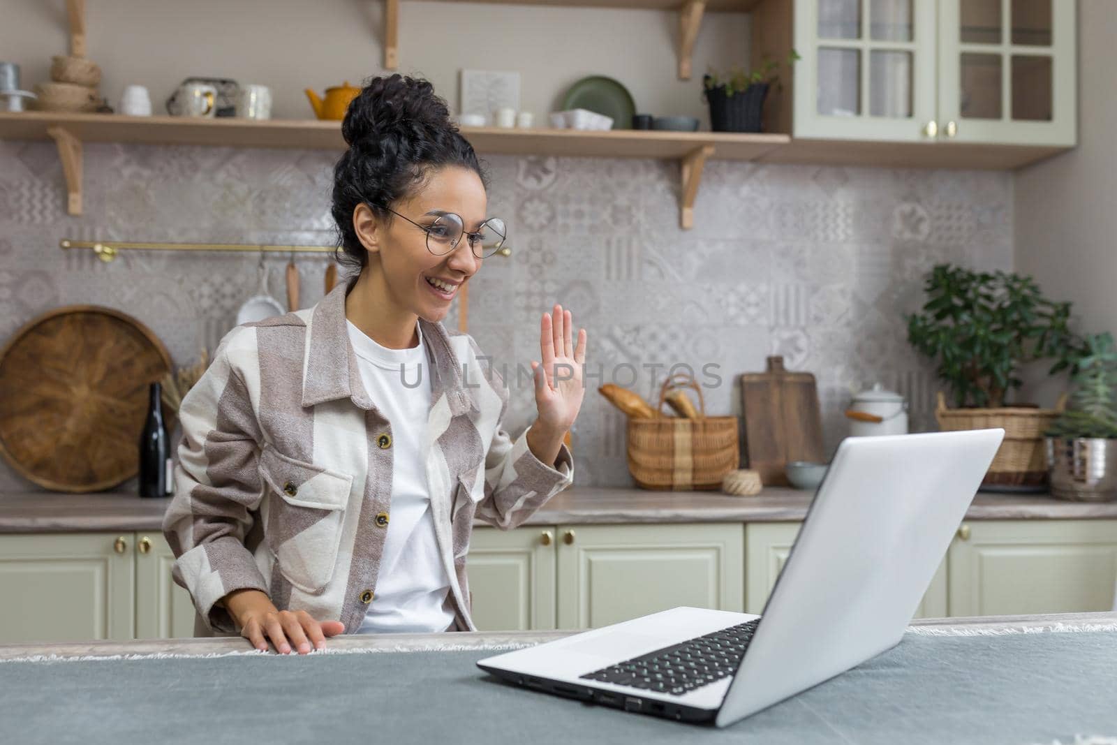 Video call with friends, young latin american woman at home sitting in kitchen waving at web camera and smiling at interlocutor, woman using laptop for remote communication.