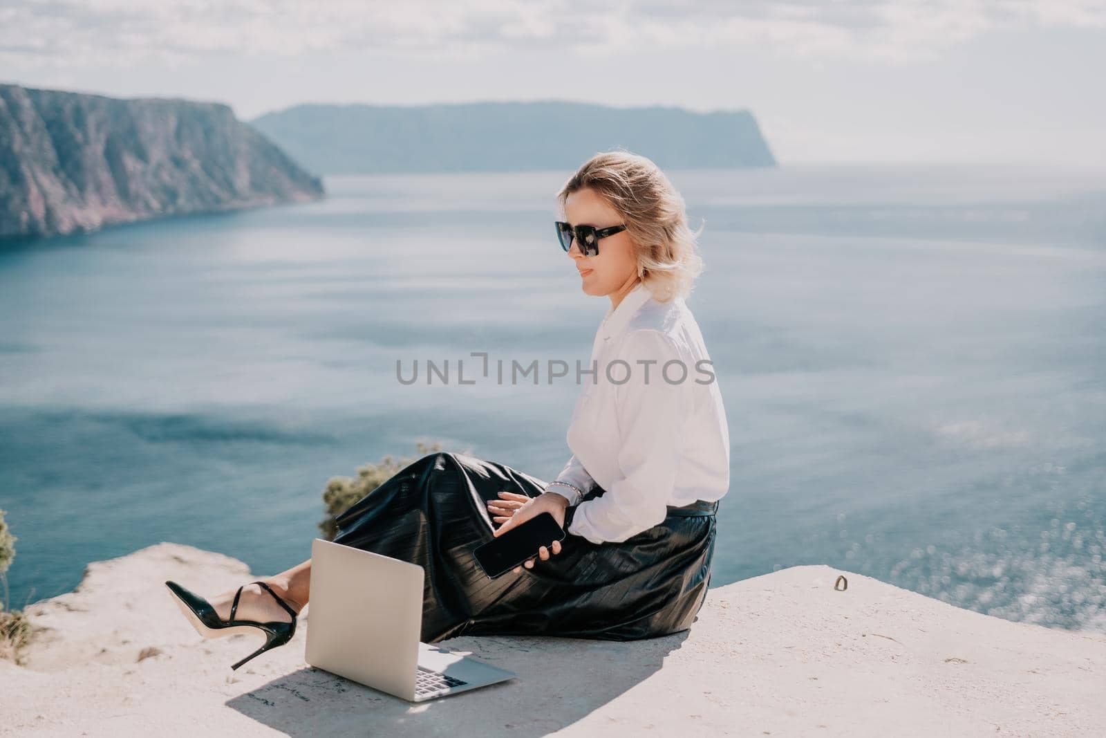 Successful business woman in yellow hat working on laptop by the sea. Pretty lady typing on computer at summer day outdoors. Freelance, travel and holidays concept.