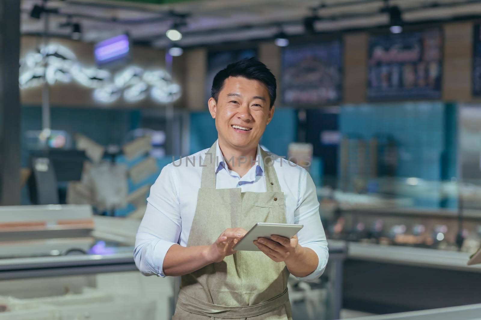 Portrait of a handsome young Asian man, an employee of a store, supermarket. Standing in an apron, holding a notepad, looking at the camera, smiling