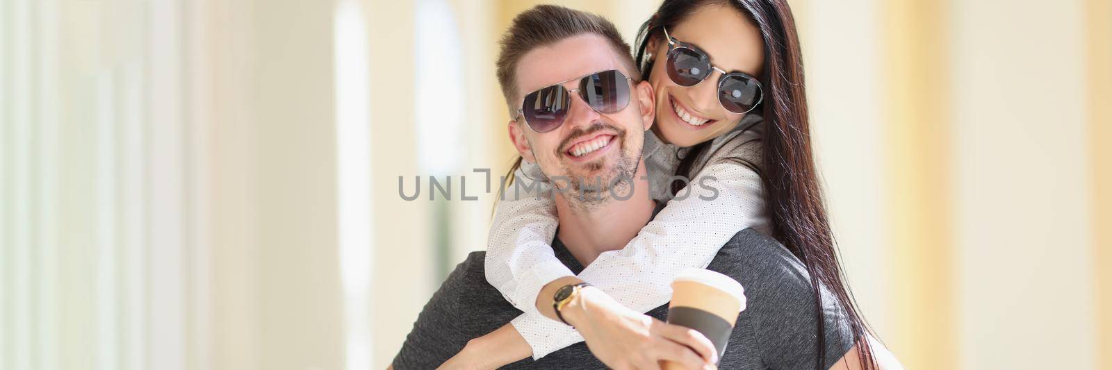 Portrait of happy couple posing, woman jumped on man back hold cup of coffee. Cheerful married people on vacation. Love, relationship, happiness concept