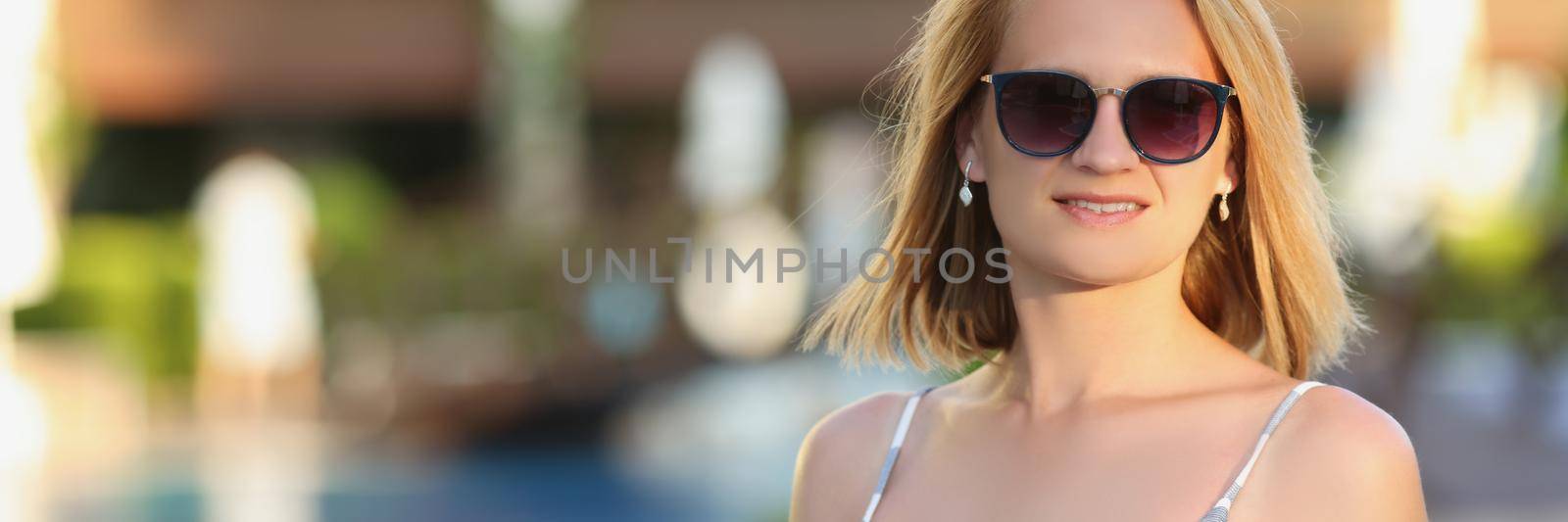 Portrait of lovely woman walking on hotel territory alone on summer evening. Blonde female in dress with resort on background. Holiday concept. Copy space