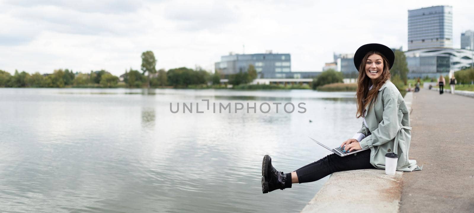 cheerful stylish woman freelancer working online on the embankment with a laptop in her hands dangling her legs to the water.