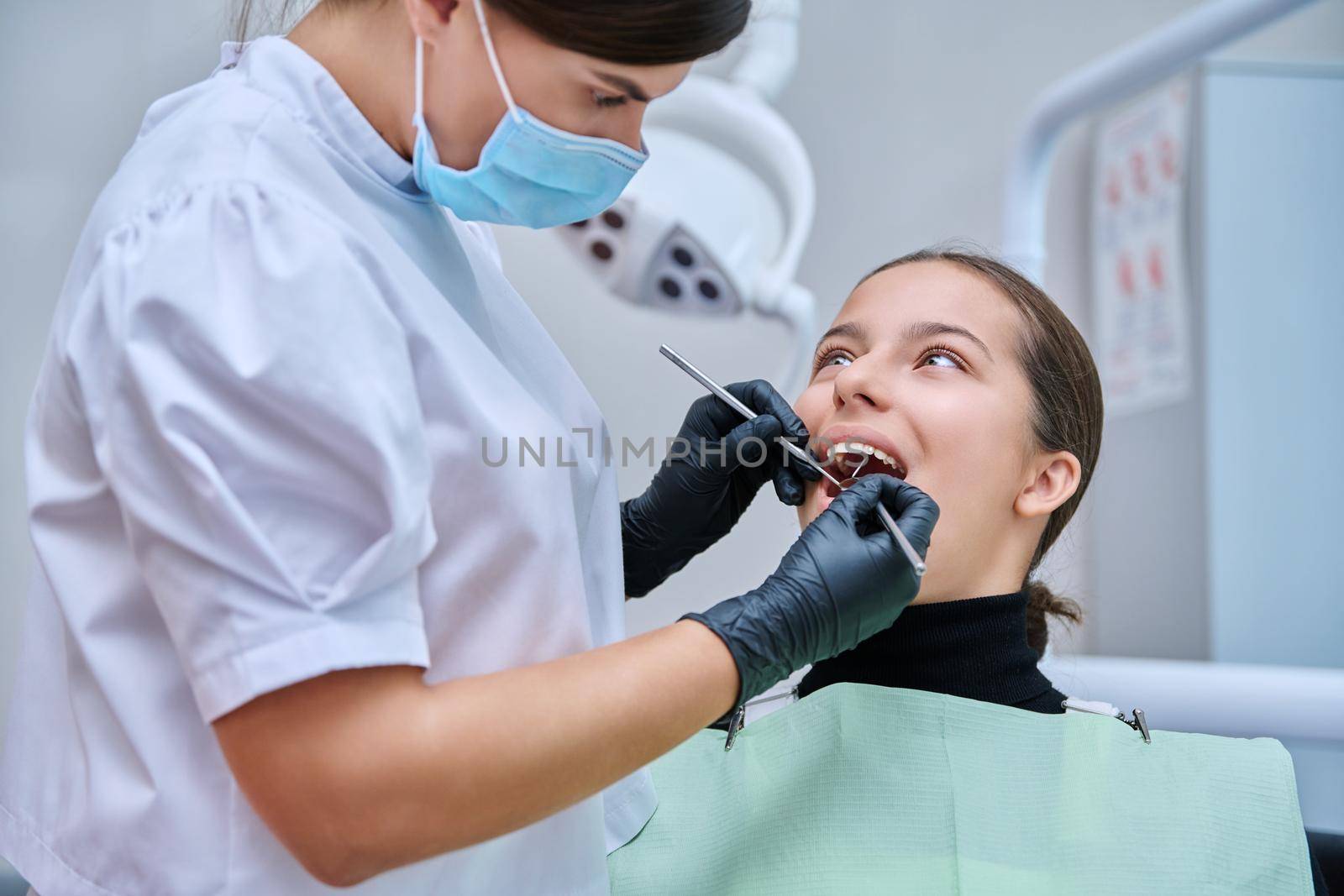 Young teenage female at dental checkup in clinic. Teenage girl sitting in chair, doctor dentist with tools examining patient's teeth. Adolescence, hygiene, treatment, dental health care