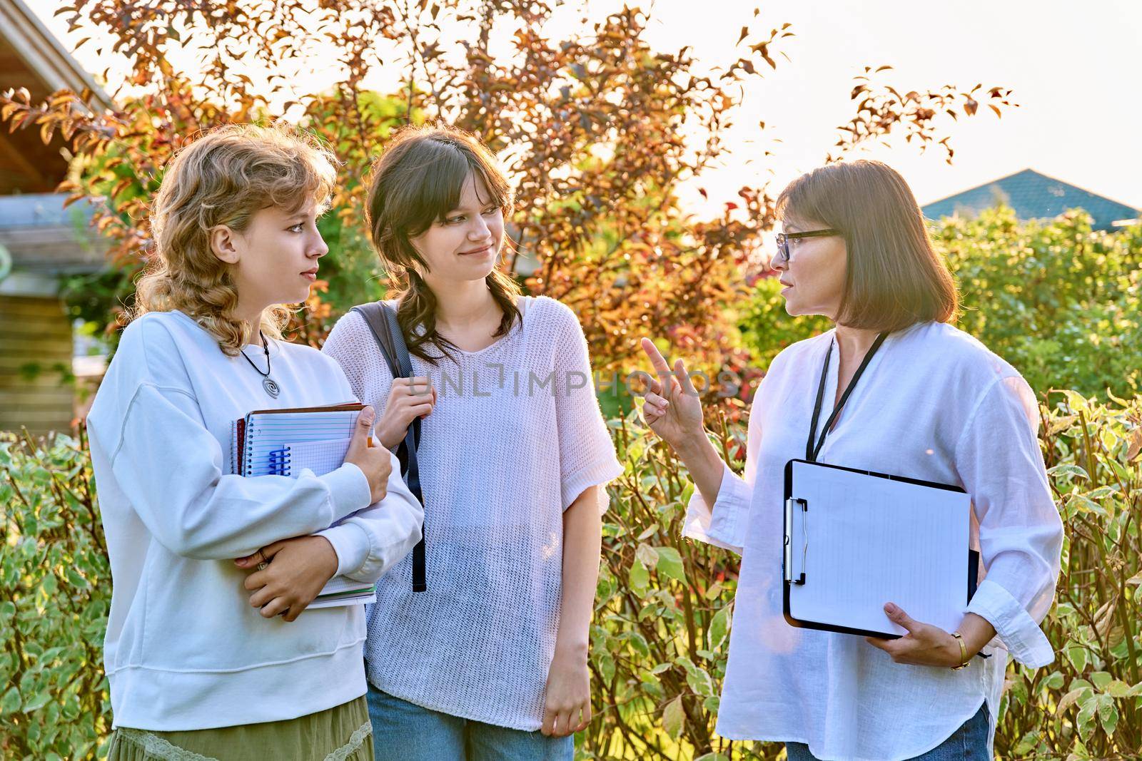 Female teacher talking to teenage female students outdoor. Two girls 17, 18 years old with middle age mentor. Teaching, education, youth, high school, college, university concept