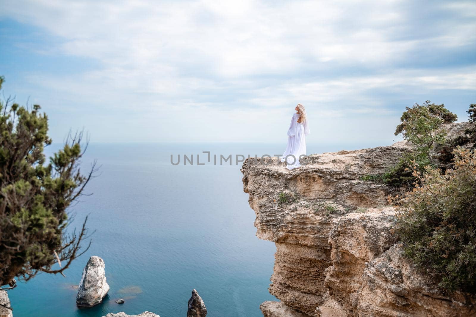 Blonde with long hair on a sunny seashore in a white flowing dress, rear view, silk fabric waving in the wind. Against the backdrop of the blue sky and mountains on the seashore