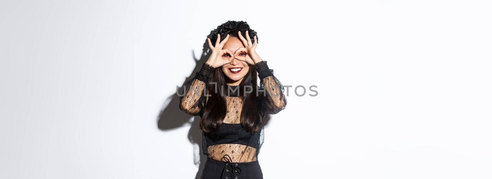 Beautiful asian woman in witch costume looking through finger glasses and smiling happy, standing over white background.