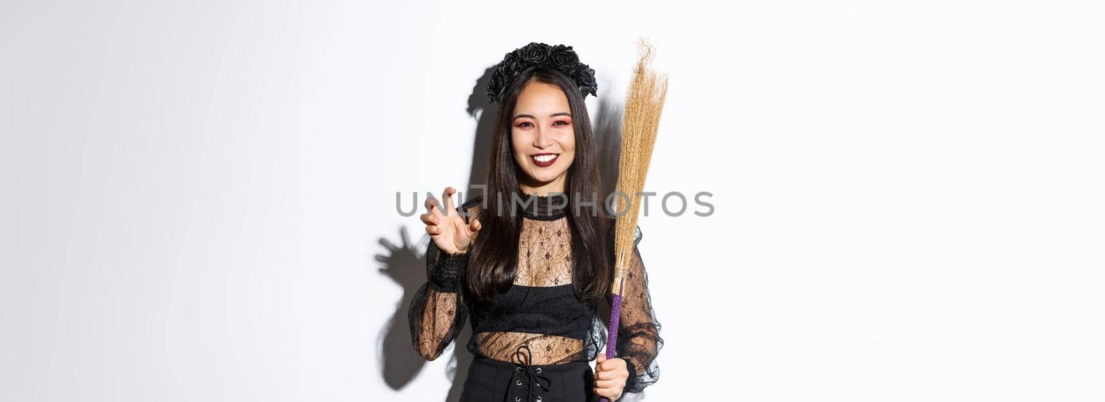 Image of beautiful asian woman in witch costume holding broom and smiling at camera, celebrating halloween, standing over white background by Benzoix