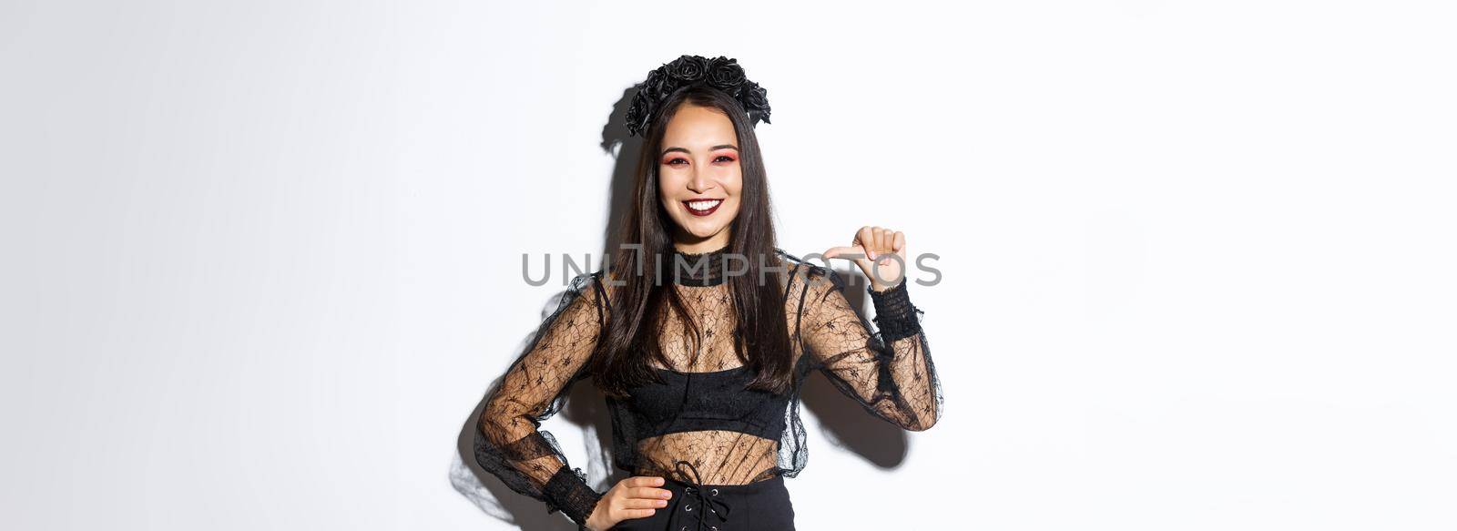 Confident smiling woman feeling like professional, pointing at herself while standing over white background in black gothic dress for halloween party.