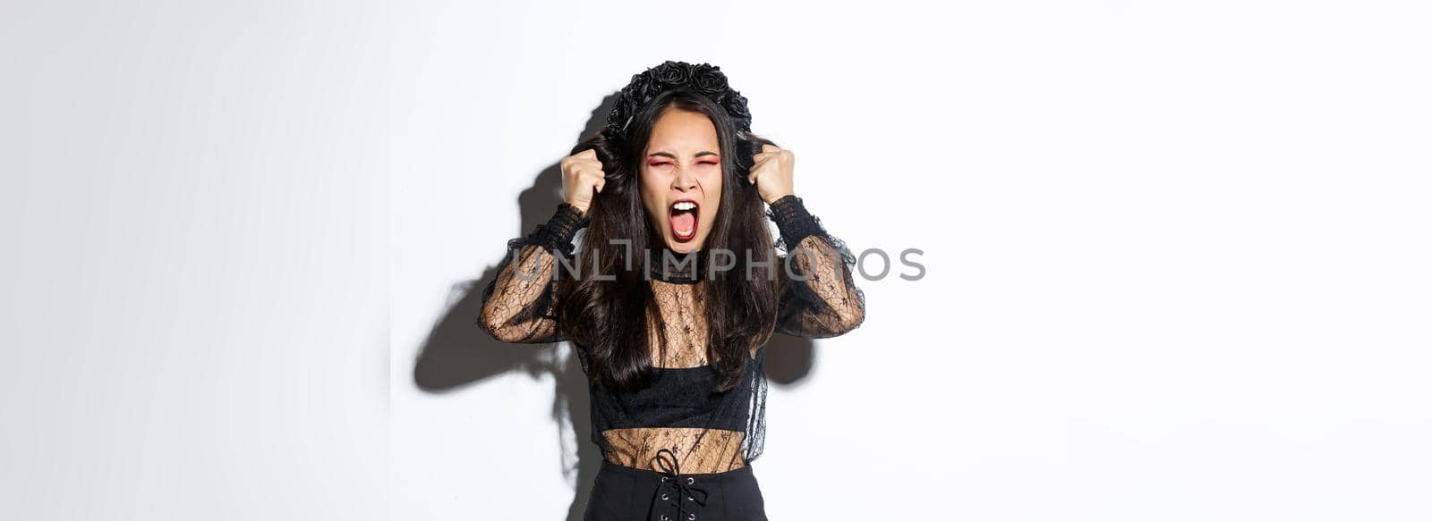 Image of angry distressed asian woman screaming and ripping hair of head, losing temper, standing in halloween costume over white background.