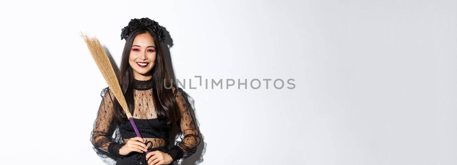 Portrait of smiling beautiful asian woman in witch costume holding broom and looking happy at camera, celebrating halloween, enjoying trick or treating, white background by Benzoix