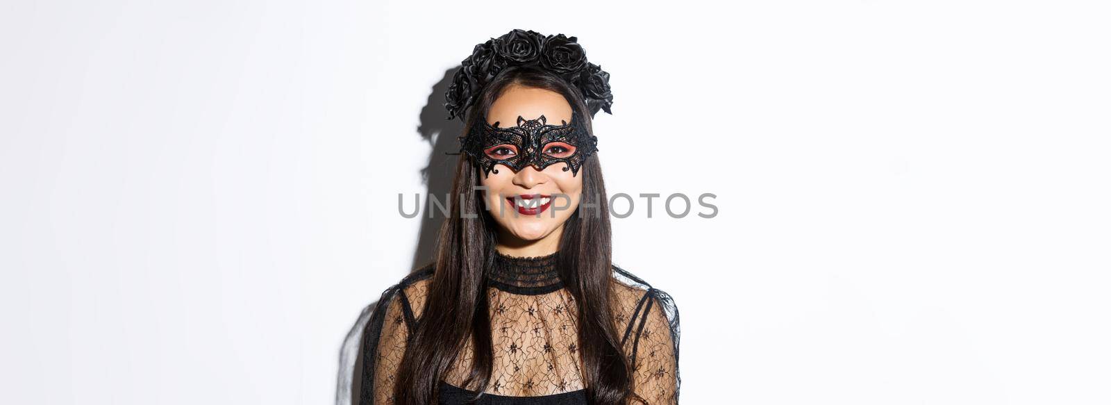 Close-up of mysterious woman in gothic wreath and black mask smiling at camera, celebrating halloween, standing over white background by Benzoix