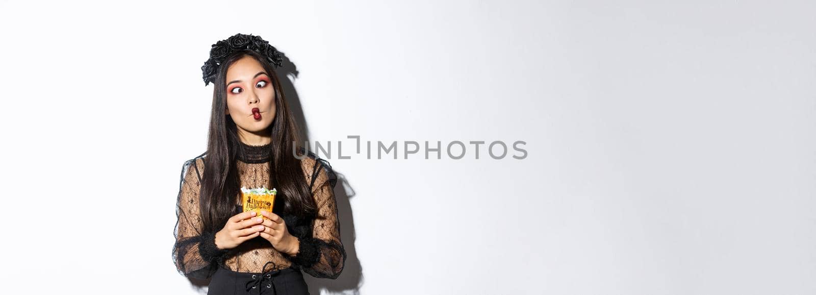 Image of funny carefree girl celebrating halloween in witch costume, making silly faces and holding treats, standing over white background.