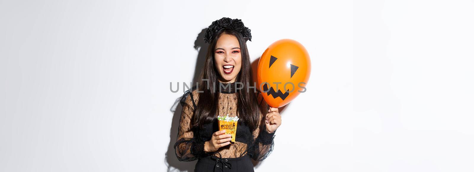 Scary witch trick or treating on halloween, holding sweets and orange balloon, standing in gothic lace dress with black wreath.