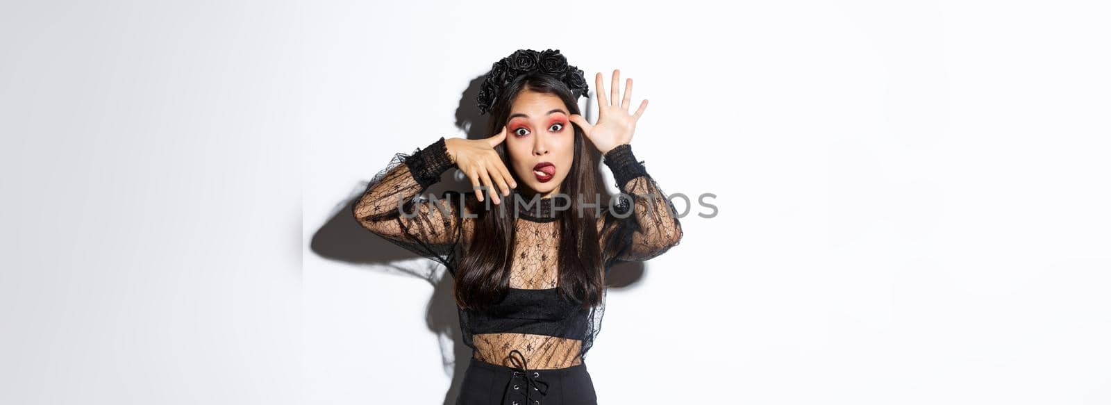 Portrait of funny asian woman in witch costume celebrating halloween, mocking someone, showing faces and sticking tongue, standing over white background.