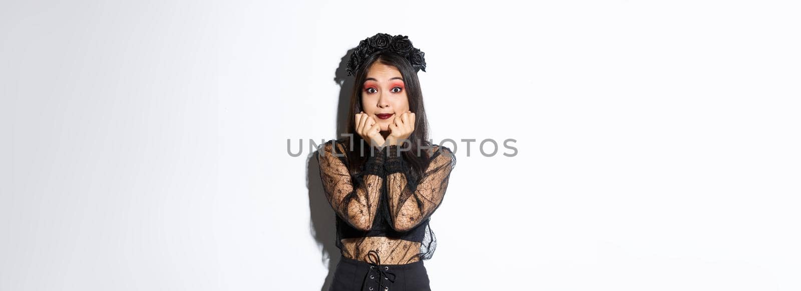 Excited cute asian girl in witch costume looking hopeful at camera, daydreaming while standing over white background, wearing gothic lace dress with wreath by Benzoix