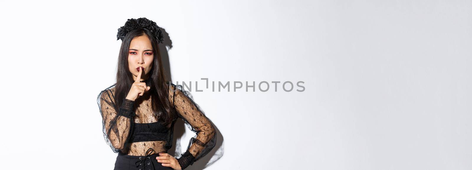 Mysterious asian witch shushing at you with concerned face, hiding secret, tell be quiet. Woman in undead widow costume prepare surprise for halloween, standing over white background.