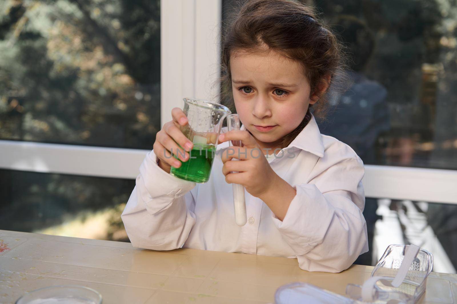 Caucasian little girl in white lab coat, conducts chemical experiment, watching a reactivity taking place in test tube with citric acid and soda, in the school chemistry laboratory. Volcanic eruption