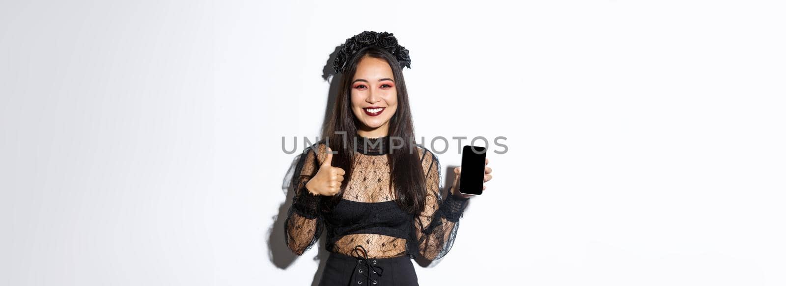 Image of happy and satisfied asian woman in halloween costume showing thumbs-up and demonstrating mobile phone screen, smiling pleased, standing over white background.